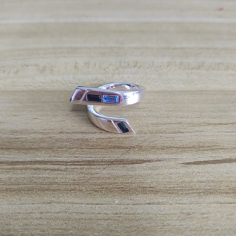 A silver-toned wrap ring featuring three small rectangular inlay stones in blue, red, and dark green, displayed on a light wood grain surface.