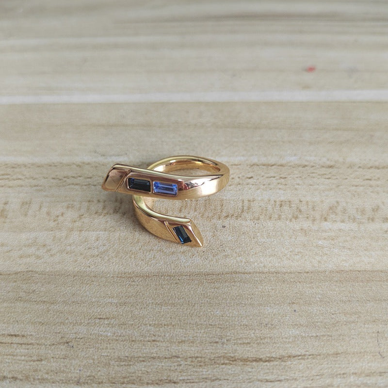 A gold wrap ring with small rectangular blue and black inlay stones, resting on a light wood surface.