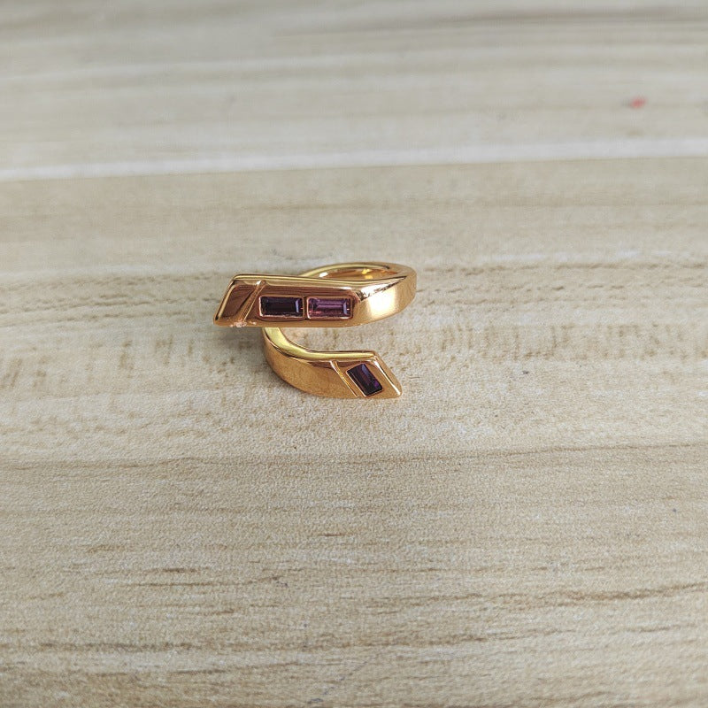 A gold-toned wrap ring with small rectangular dark red gemstone accents, resting on a light wood surface.