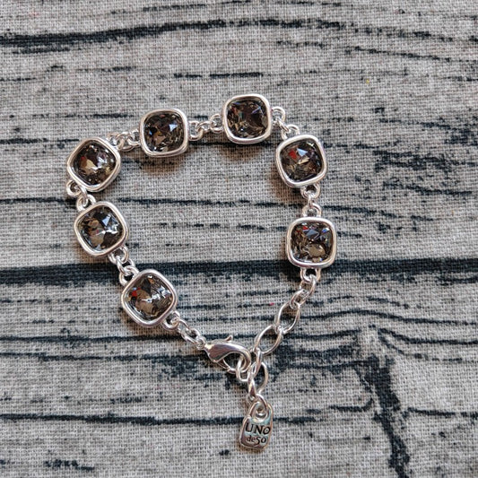 A silver chain bracelet with seven cushion-cut smoky brown gemstones, displayed on a textured gray fabric background. A small hangtag engraved with 'UNO 16K' is attached to the clasp.