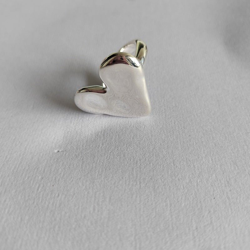 A polished silver heart-shaped ring with a smooth, engraved surface and a polished band, resting on a textured light gray paper background.