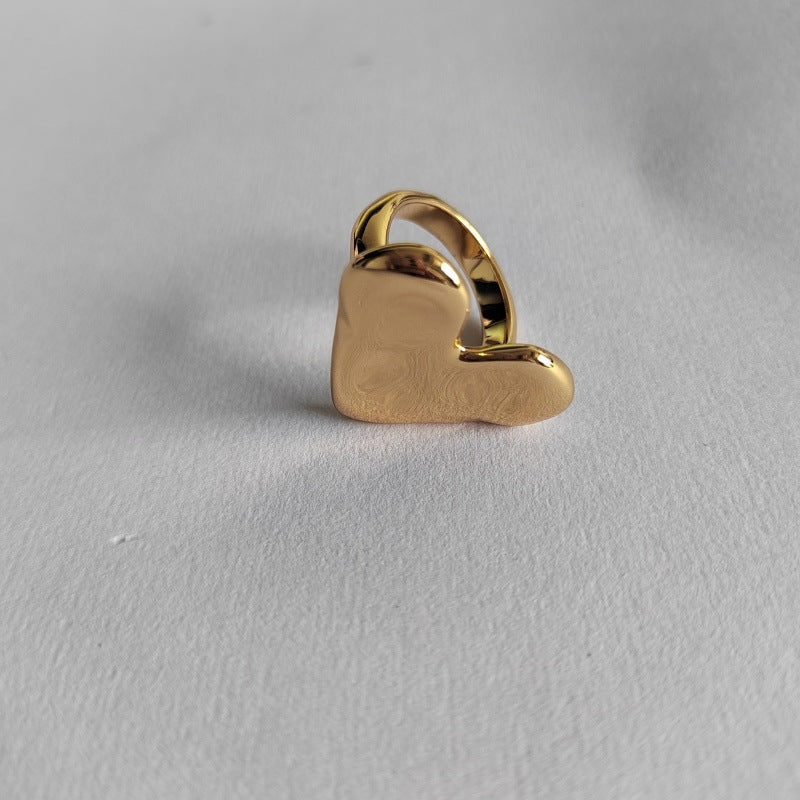 A polished gold heart-shaped ring with a smooth, reflective finish, displayed on a textured light gray surface.