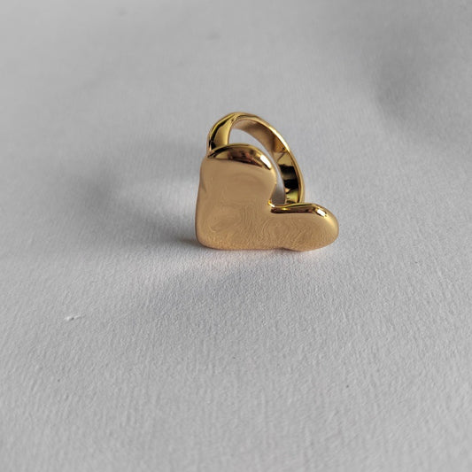 A polished gold heart-shaped ring with a smooth, reflective finish, displayed on a textured light gray surface.