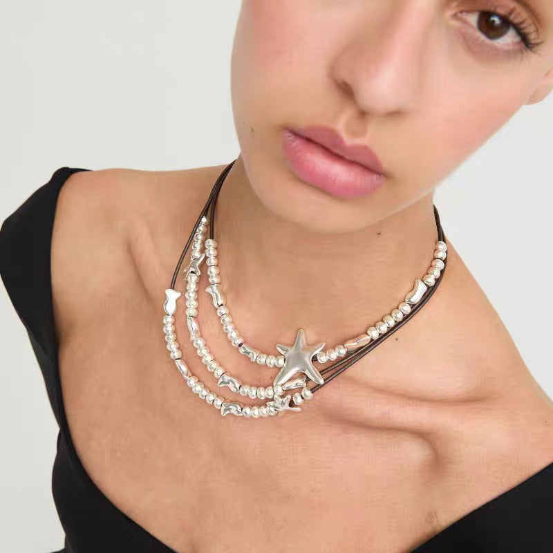 A close-up of a model wearing a multi-strand necklace featuring white faux pearls, silver starfish charms, and small silver shells on dark cords, paired with a black off-the-shoulder top.