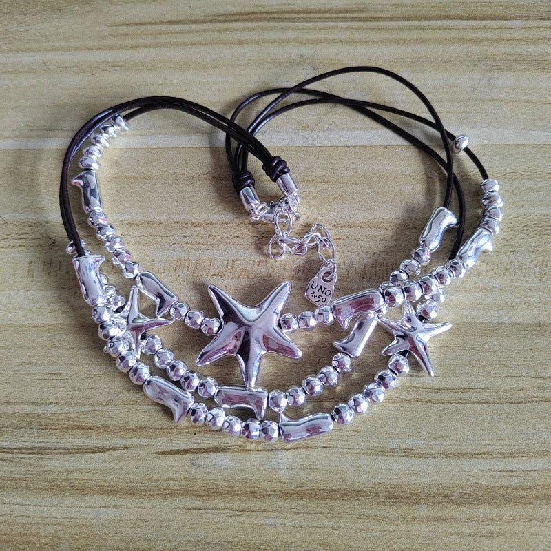 A set of three layered bracelets featuring silver starfish charms, silver beads, and dolphin charms on black cords, displayed on a light wood background.
