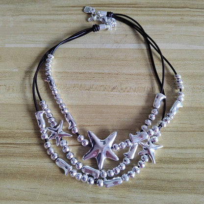 A multi-strand necklace featuring silver starfish charms, silver beads, and silver shell-shaped beads on black cords, displayed on a light wood surface.