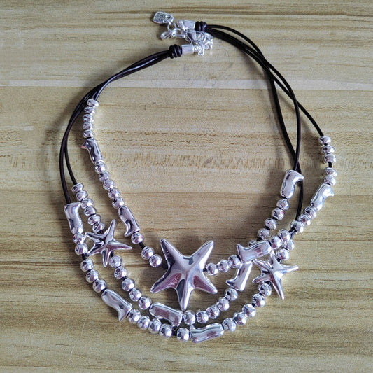 A multi-strand necklace featuring silver starfish charms, silver beads, and silver shell-shaped beads on black cords, displayed on a light wood surface.