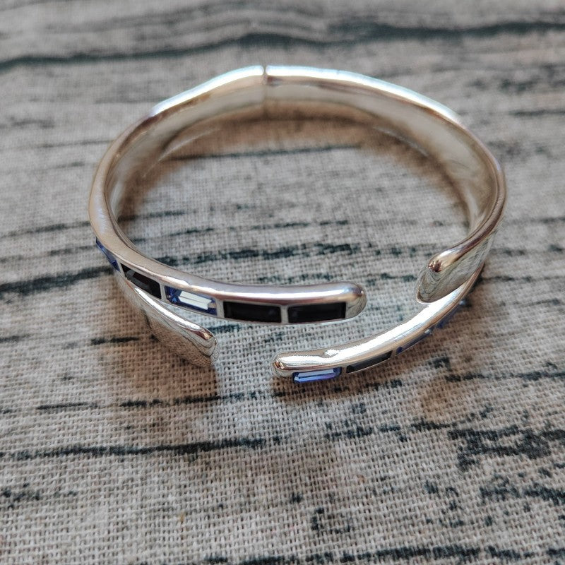 A polished silver-toned cuff bracelet with inlaid black and blue rectangular accents, photographed on a textured gray fabric background.