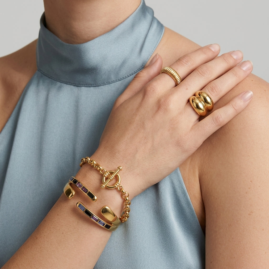 A close-up of a person wearing multiple gold jewelry pieces, including a chain bracelet with colorful inlay, a ring, and a bangle, paired with a light blue high-neck top against a neutral gray background.