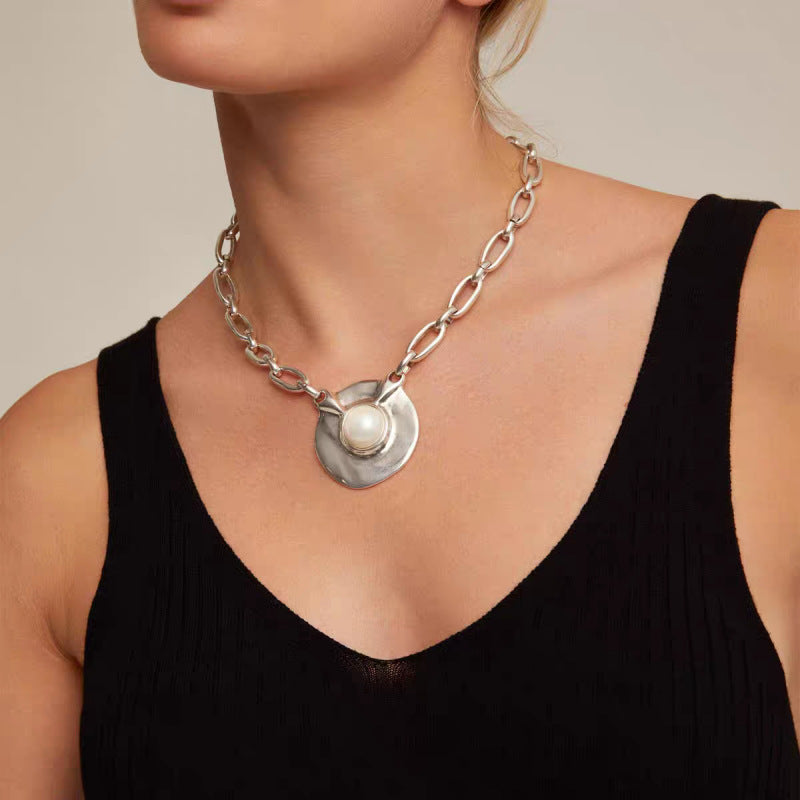 A close-up of a model wearing a chunky silver chain necklace with a textured circular pendant featuring a central white pearl, paired with a black ribbed tank top.