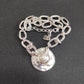 A silver chain bracelet with a hammered round disc pendant and a central white pearl, displayed on a dark textured background.