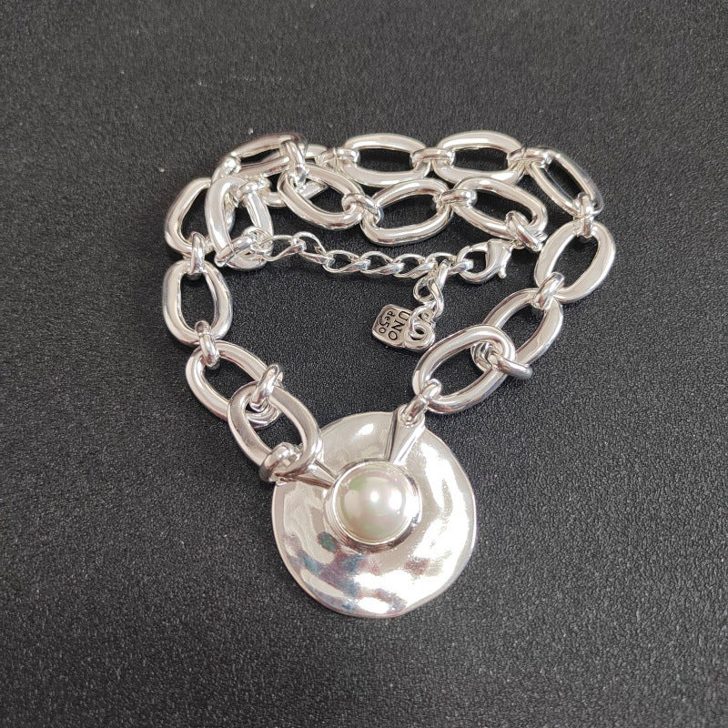 A silver chain bracelet with a hammered round disc pendant and a central white pearl, displayed on a dark textured background.