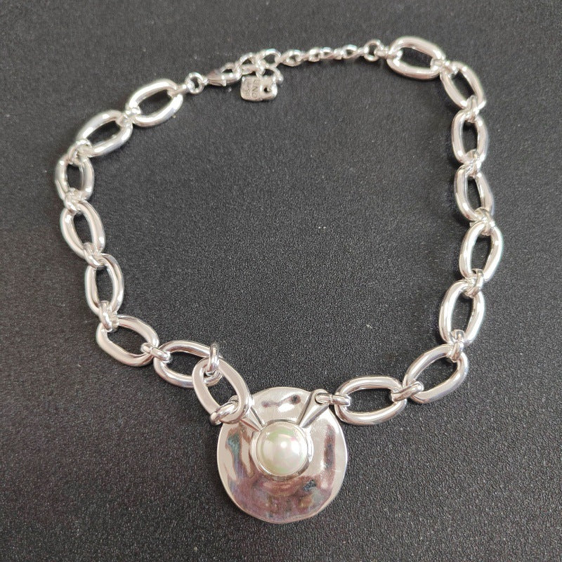 A silver chain bracelet with elongated oval links and a central hammered silver disc featuring a white pearl, displayed on a dark textured background.