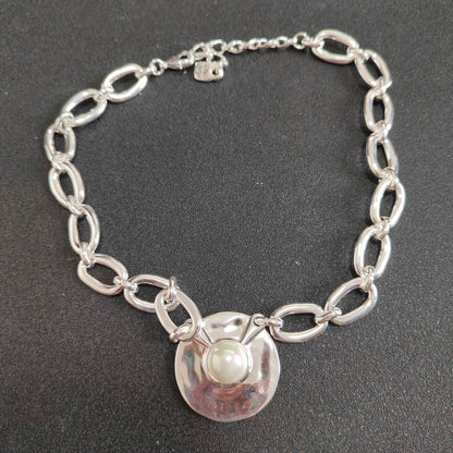 A silver chain bracelet with elongated oval links and a central hammered silver disc featuring a white pearl, displayed on a dark textured background.