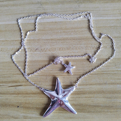 A silver-toned necklace featuring a large polished starfish pendant and a smaller starfish charm, displayed on a light wood grain surface.