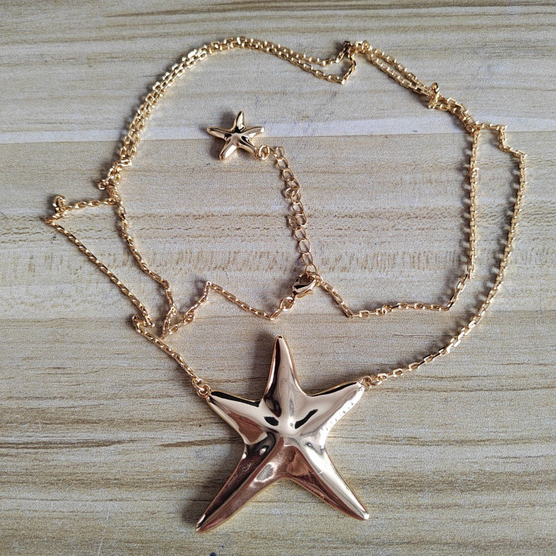 A gold-toned necklace featuring a large starfish pendant and a smaller matching starfish charm, displayed on a light wood grain surface.