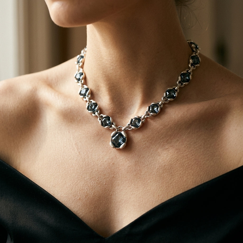 A close-up of a model wearing a silver-toned necklace with dark blue-gray marquise-cut gemstones, paired with a black off-the-shoulder top.