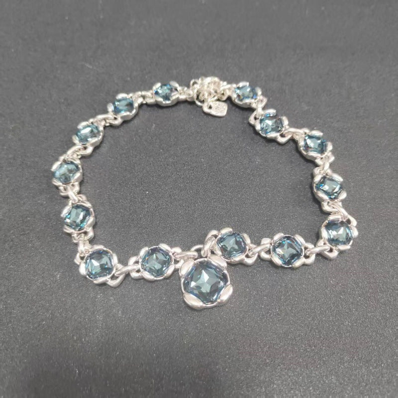 A silver-tone bracelet featuring a series of faceted light blue gemstones in a prong setting, with a larger matching blue stone at the center and a small heart-shaped charm near the clasp, displayed on a dark gray textured background.