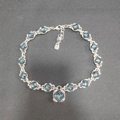 A silver-tone bracelet featuring a series of round light blue faceted gemstones in a floral setting, with a larger matching blue gemstone at the center and an adjustable clasp with a small tag, displayed on a dark gray textured background.