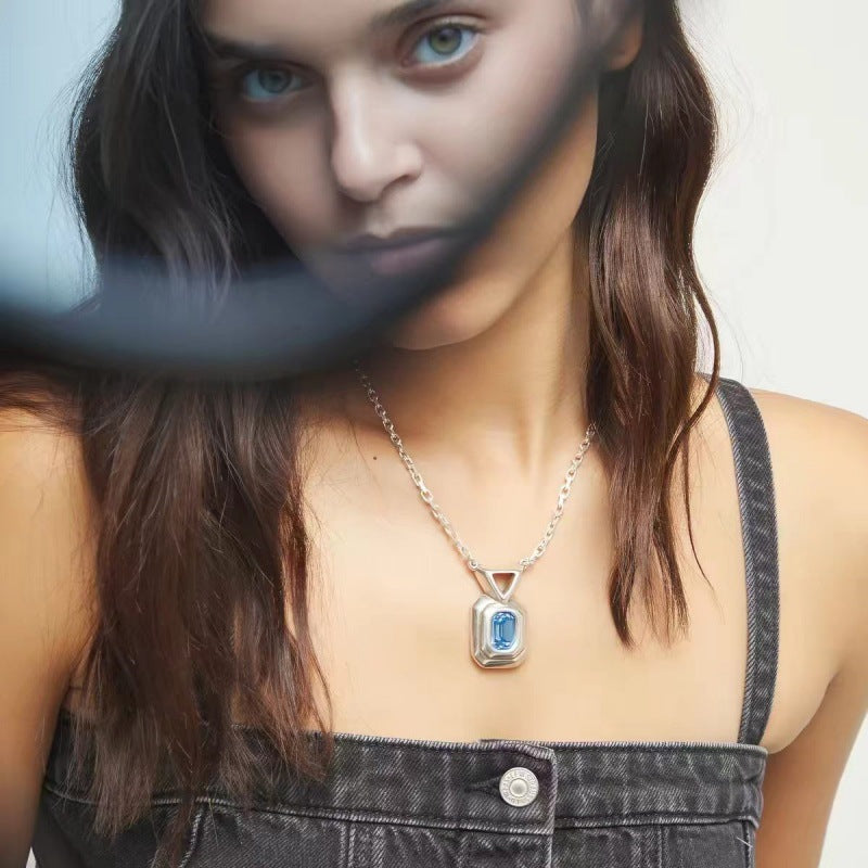 A close-up lifestyle shot of a model wearing a silver pendant necklace with a rectangular blue gemstone. The image has a stylized, high-contrast editing look with a dark, blurred overlay on the left side of the model's face.