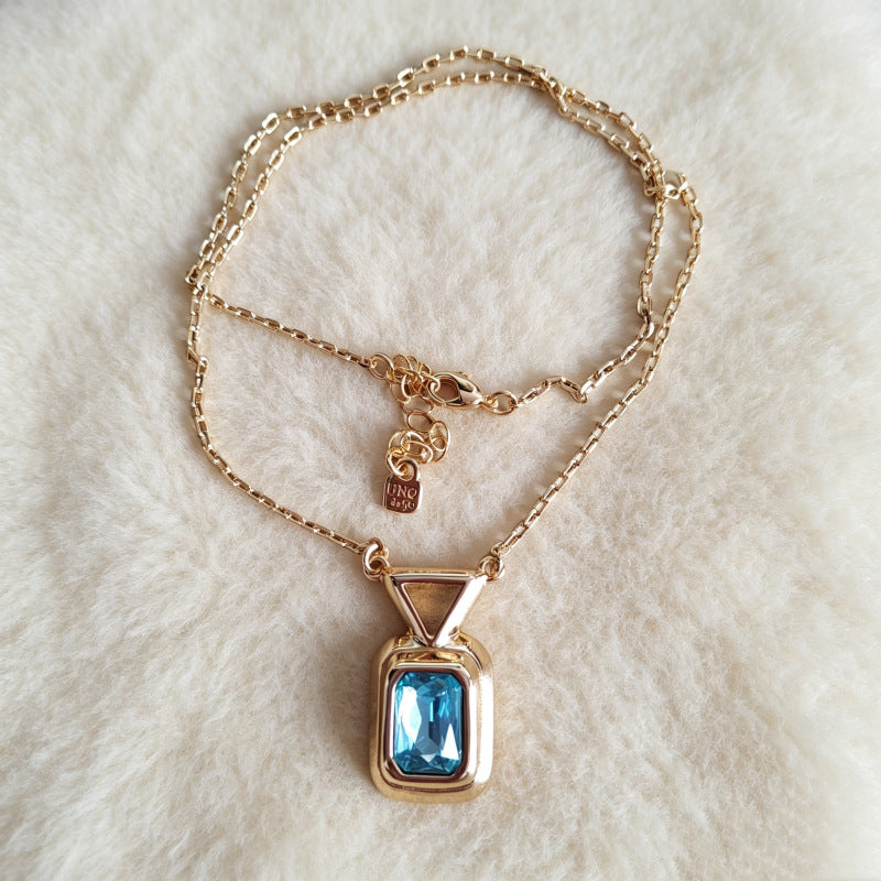 A gold chain necklace with a rectangular blue gemstone pendant, displayed on a soft white faux fur background.