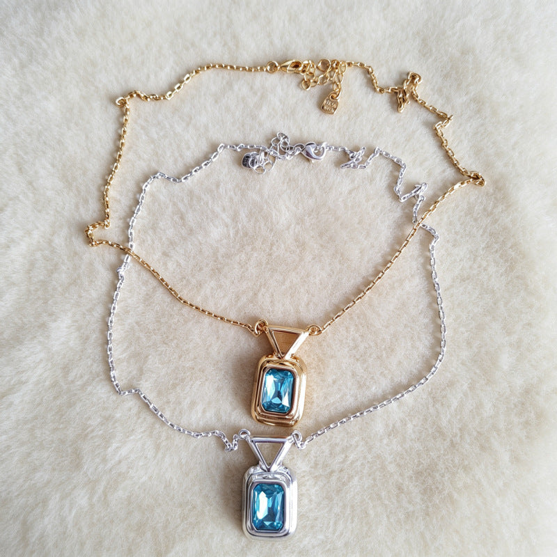 Two delicate necklaces with rectangular blue gemstone pendants, one in gold-tone metal and one in silver-tone metal, displayed on a soft white faux fur background.