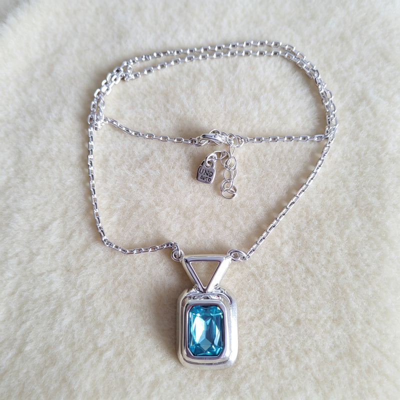 A silver chain necklace with a rectangular bezel pendant featuring a light blue gemstone, displayed on a soft cream-colored textured background. A small brand tag is attached to the clasp.