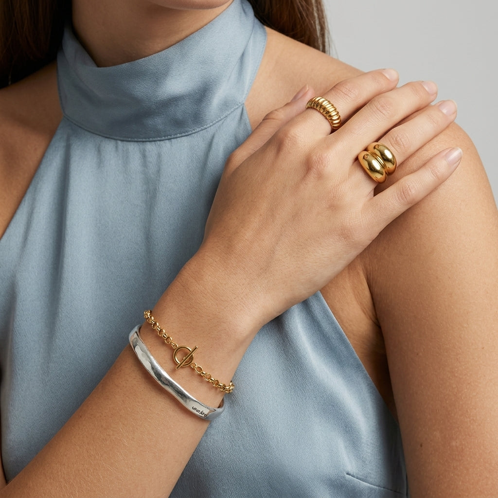A close-up of a model's hand and wrist wearing gold and silver bracelets and rings, paired with a light blue satin high-neck top against a neutral gray background.