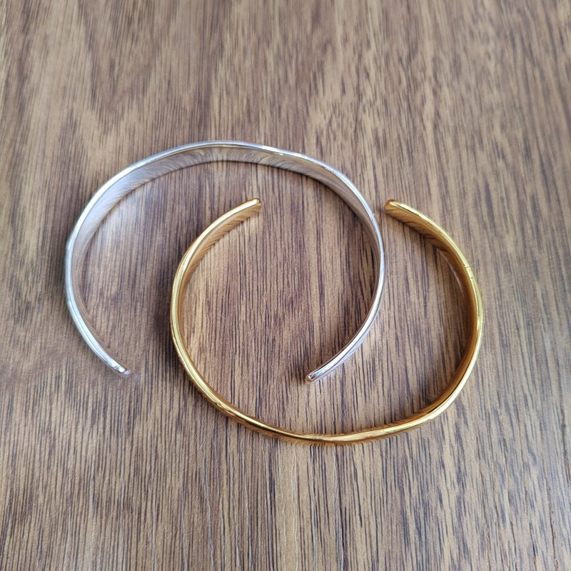 Two minimalist open cuff bracelets, one silver-toned and one gold-toned, displayed on a brown wooden surface.