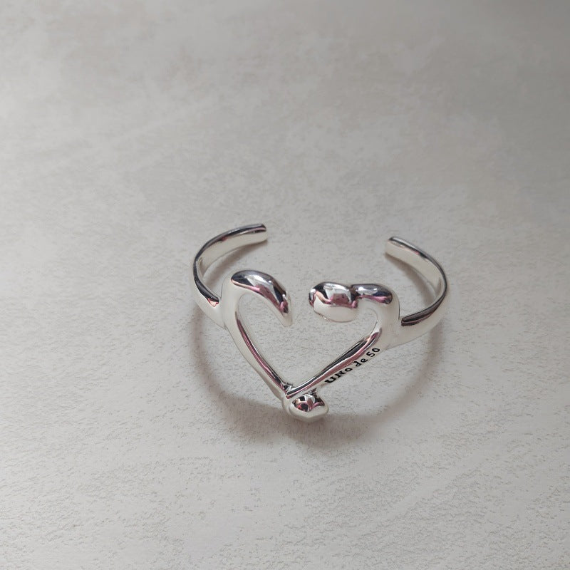 A polished sterling silver open cuff ring shaped like a heart, with the word 'LOVE' engraved on the inner band, resting on a textured light gray surface.
