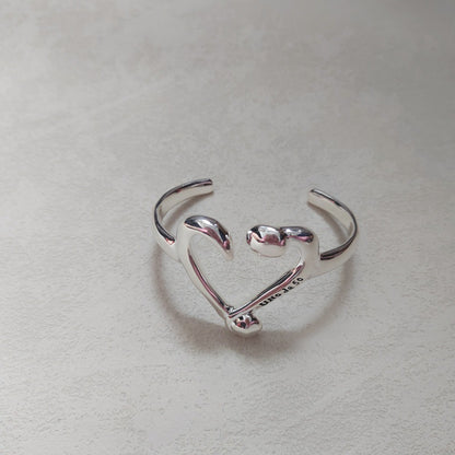 A polished sterling silver open cuff ring shaped like a heart, with the word 'LOVE' engraved on the inner band, resting on a textured light gray surface.