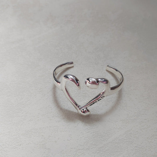 A polished sterling silver open cuff ring shaped like a heart, with the word 'LOVE' engraved on the inner band, resting on a textured light gray surface.