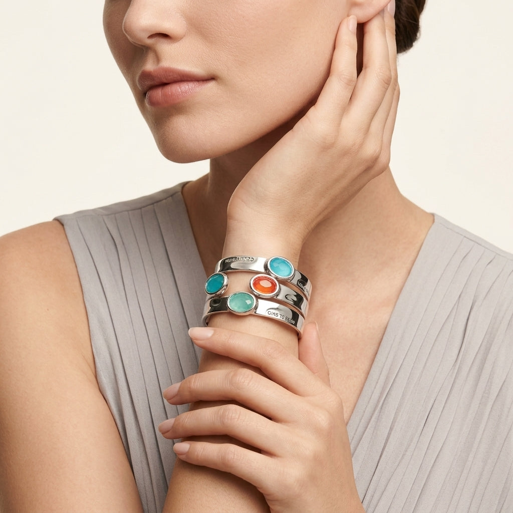 A close-up of a model wearing a stack of three silver cuff bracelets with round turquoise, green, and orange cabochon stones, paired with a light gray pleated top.