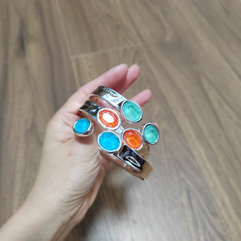 A hand holding a silver cuff bracelet with multiple oval gemstone accents in blue, orange, and green, set against a wooden floor background.