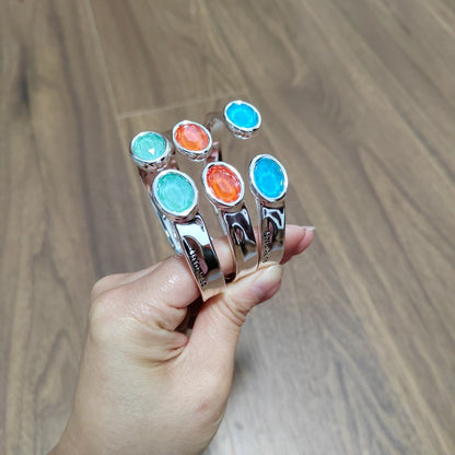 A hand holding a set of four silver cuff bracelets, each featuring a different oval gemstone in turquoise, light blue, orange, and bright blue, against a wooden background.