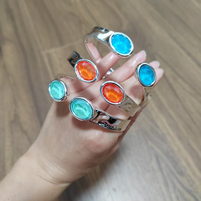 A hand wearing five silver cuff bracelets, each featuring a different oval cabochon stone in vibrant blue, orange, and mint green, set against a wooden background.