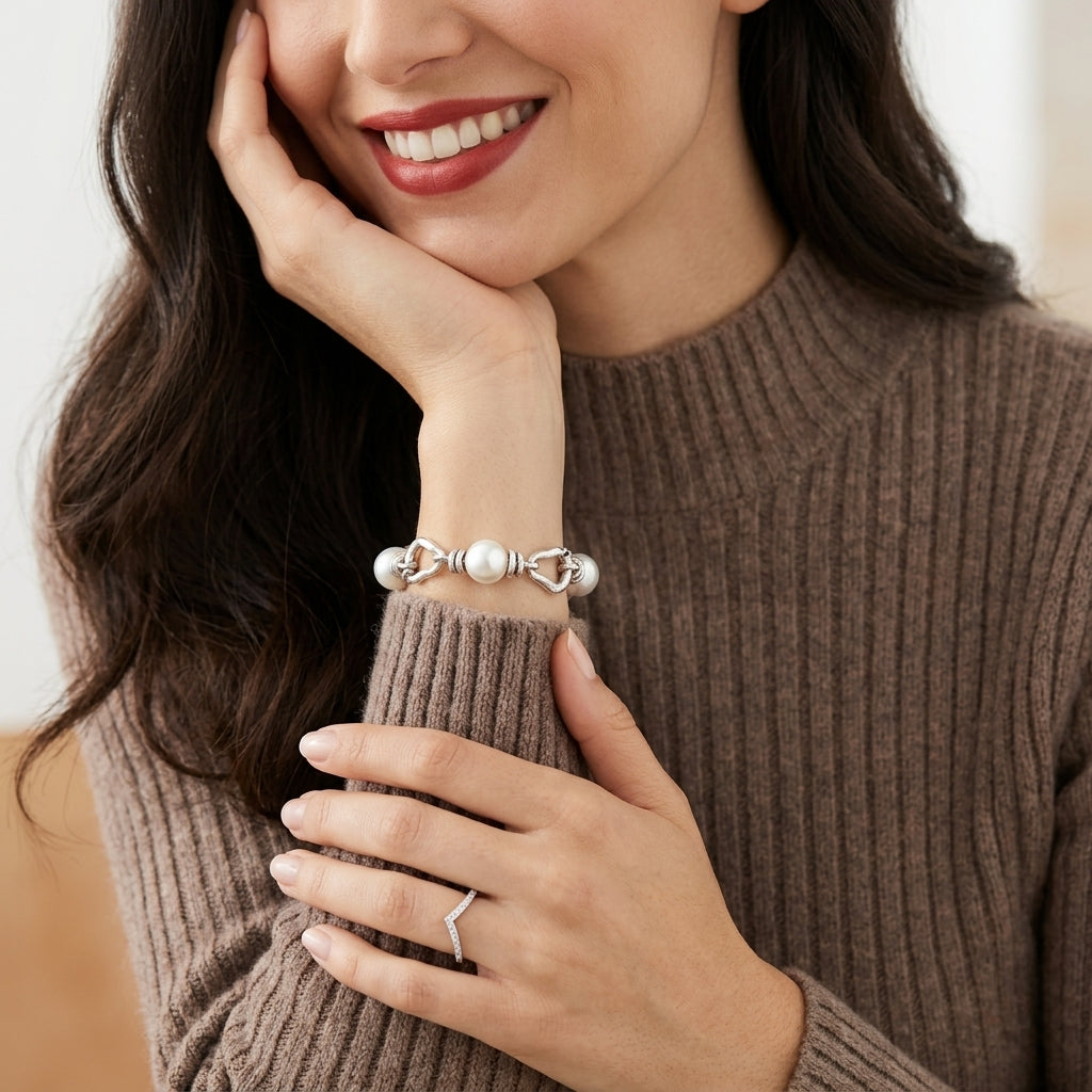 A close-up lifestyle shot of a woman wearing a silver-toned double knot bracelet with white pearl accents on her wrist, paired with a silver chevron ring on her finger. She is wearing a brown ribbed knit sweater against a soft, warm neutral background.