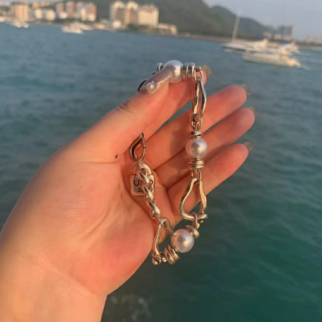 A hand holding a gold-toned chain bracelet with white pearls, set against a blurred background of blue ocean water and boats.