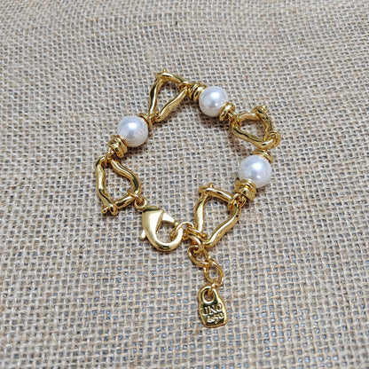 A gold-plated chain bracelet with three white pearl beads and a lobster clasp, displayed on a textured beige burlap background. A small tag engraved with 'JNDJ 925' is visible.