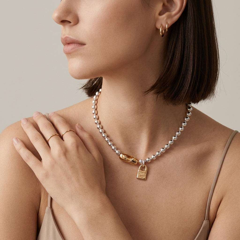 A close-up of a woman wearing a delicate pearl necklace with a gold padlock pendant, styled with gold hoop earrings and thin gold rings against a neutral beige background.