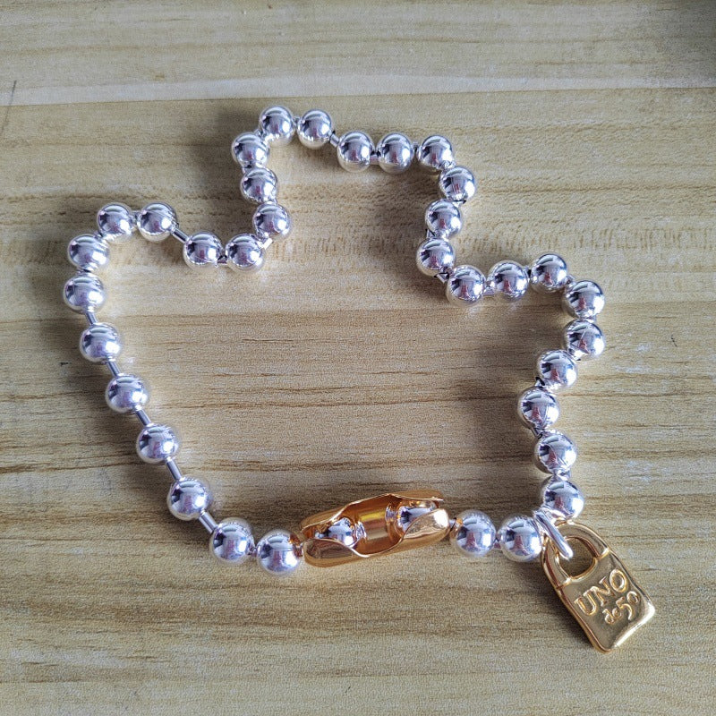 A silver beaded bracelet with a gold lobster clasp and a small gold padlock charm engraved with 'JSP', displayed on a light wood surface.