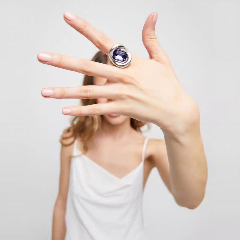 A woman wearing a large silver-toned ring with a deep purple oval gemstone, holding her hand up to show the ring against a light gray background.