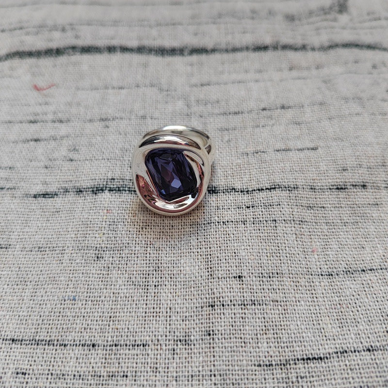 A silver-toned signet ring featuring a large, polished dark blue-purple gemstone, resting on a textured gray linen fabric background.