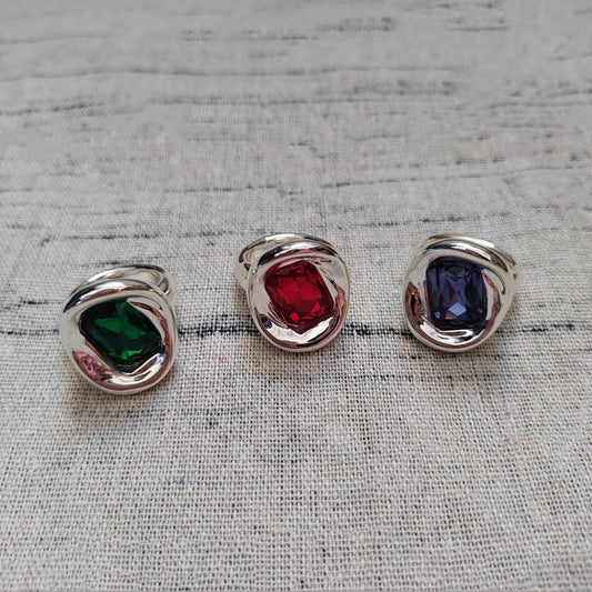 Three silver-toned ring settings with square-cut gemstones in green, red, and dark blue, arranged on a textured light gray fabric background.