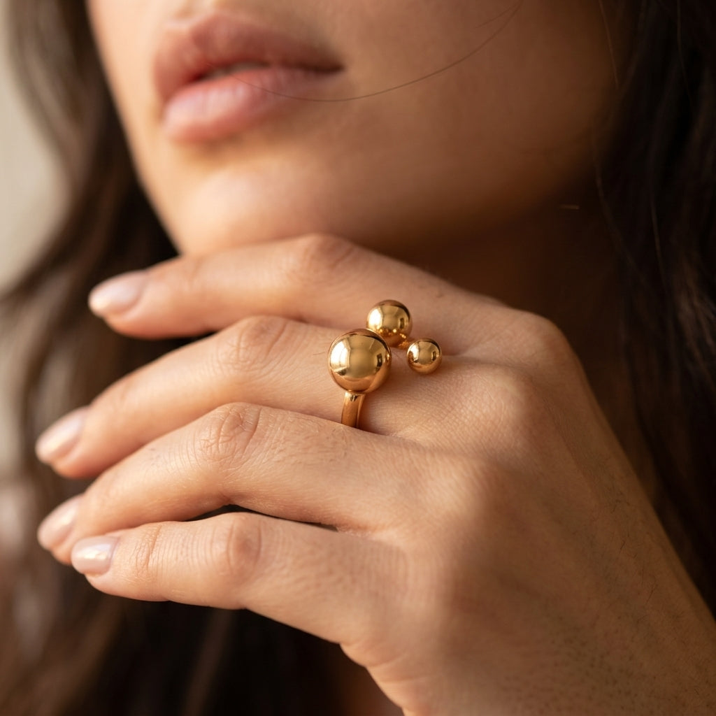 A close-up of a woman's hand wearing a gold ring with three rounded, clustered ball motifs on a soft beige background.