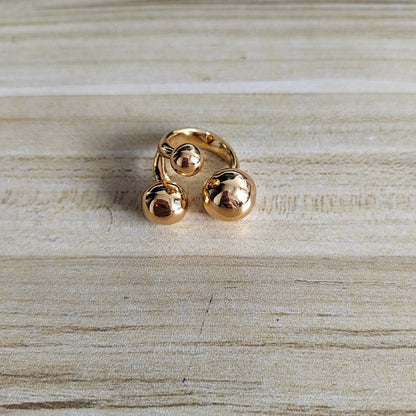 A gold-colored adjustable ring with three polished spherical beads, displayed on a light wood grain surface.