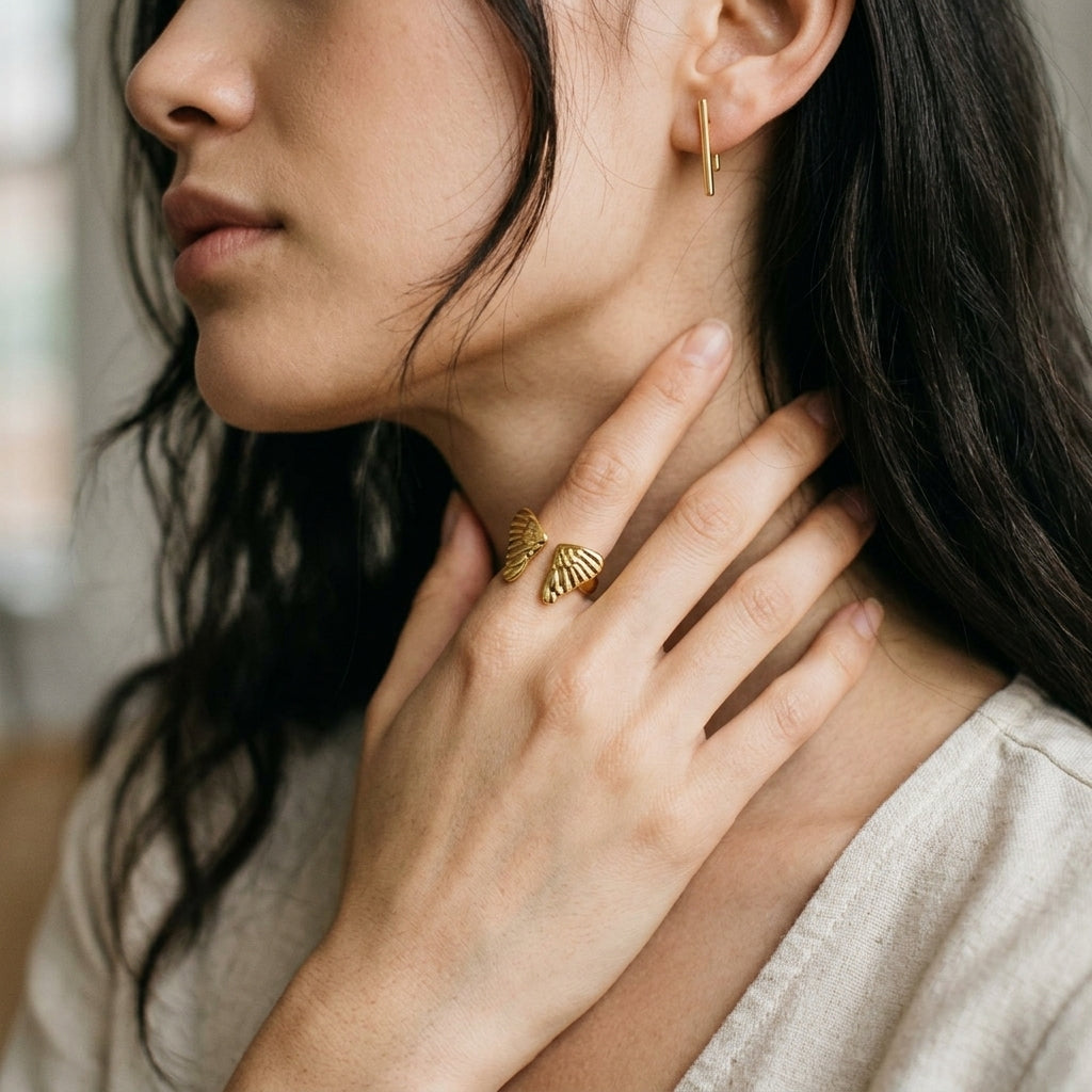 A close-up lifestyle shot of a woman wearing a delicate gold ring shaped like a butterfly on her ring finger, with a matching small gold hoop earring visible on her ear.