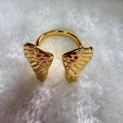 A gold-plated ring featuring two detailed angel wing motifs on either side of a circular band, displayed on a soft white faux fur surface.