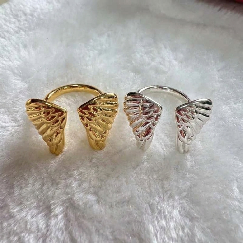 Two angel wing open-bend rings, one in yellow gold and one in silver, displayed side-by-side on a white faux fur surface.