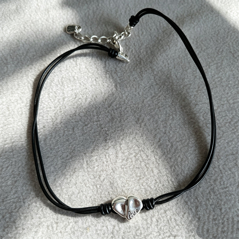 A black double-strand cord bracelet with a silver split-heart charm engraved with 'love', displayed on a textured light gray fabric under direct sunlight.